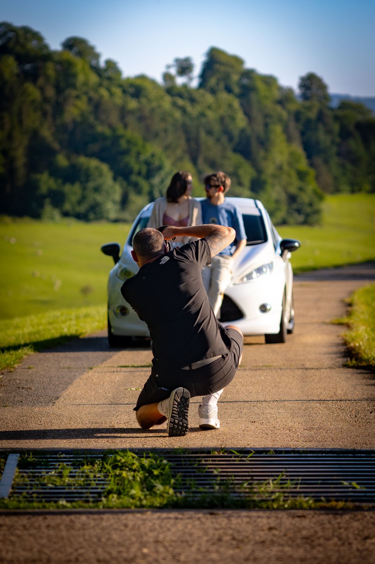 Photographer crouches low to capture shot of car on rural road with green hills in background.