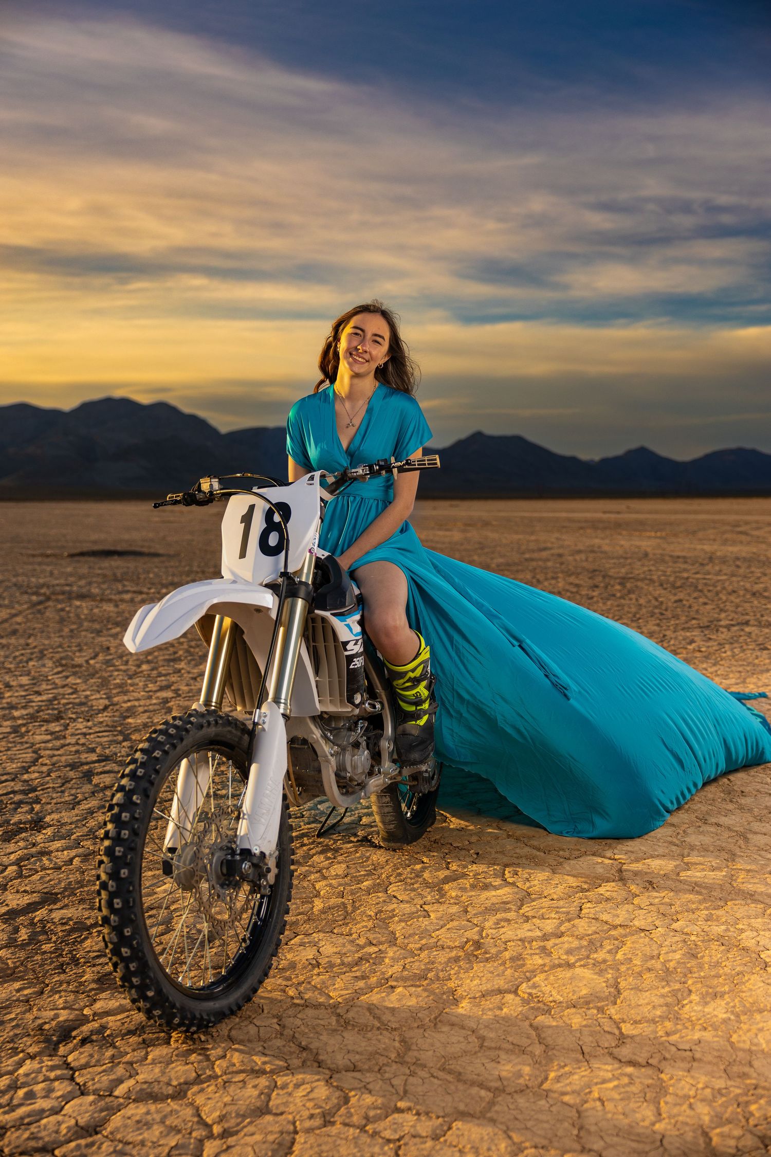 Dirt bike rider in flowing turquoise dress and yellow socks poses on dry cracked earth at sunset with mountain backdrop.