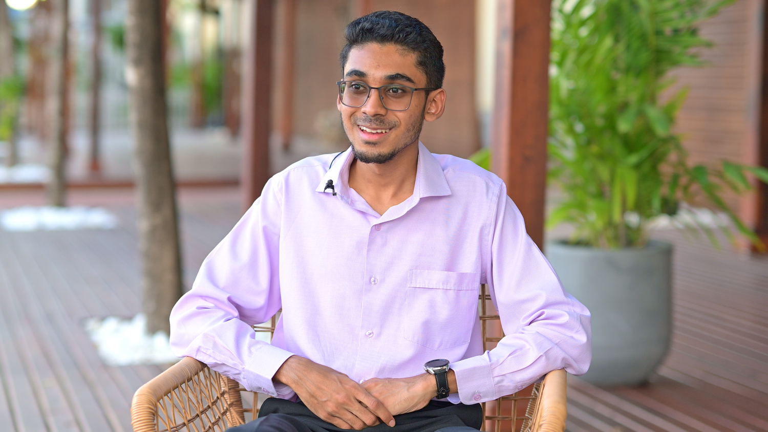 Portrait of person in lavender dress shirt seated in wicker chair against greenery backdrop.