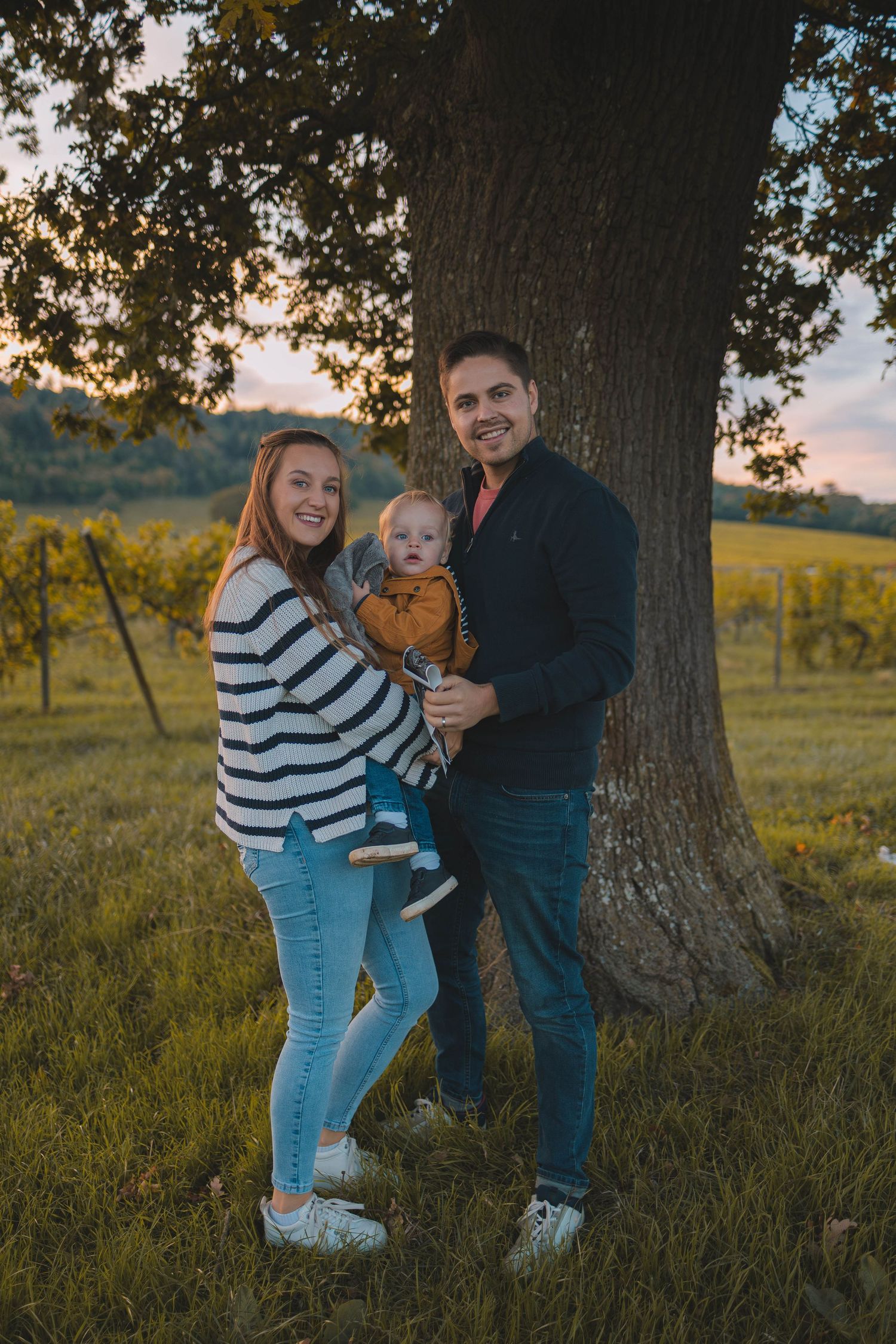 Family stands together at sunset in front of large tree in vineyard setting.