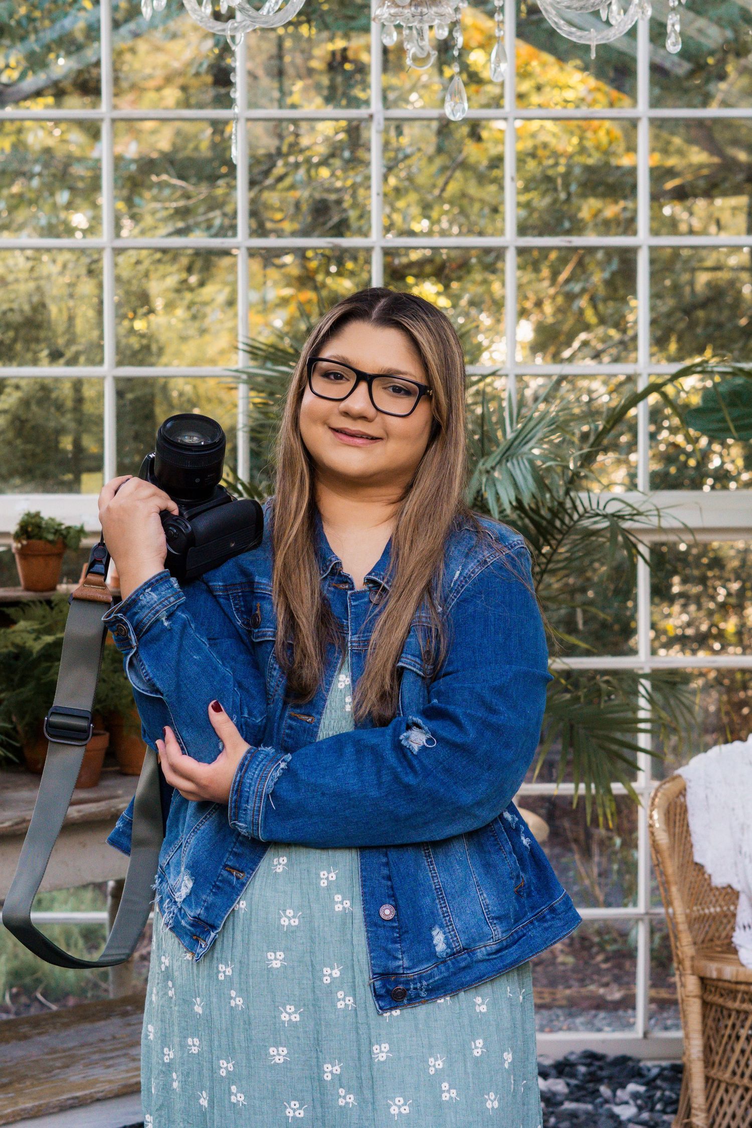 Photographer in striped sweater holds camera while posing against white wall with plants.