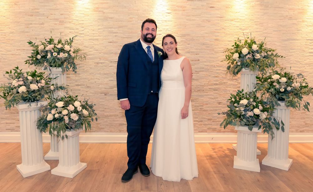 A couple poses in formal wedding attire in front of a brick wall with white floral arrangements on pedestals.