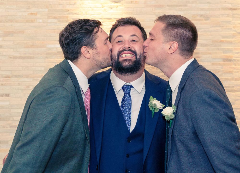 Three people in formal suits share a joyful moment at what appears to be a wedding celebration.