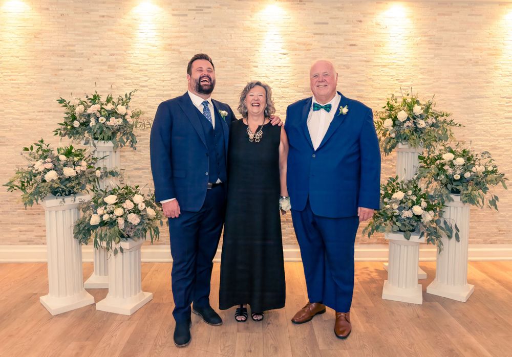 Three people in formal attire stand between floral arrangements at an elegant event venue with warm lighting.
