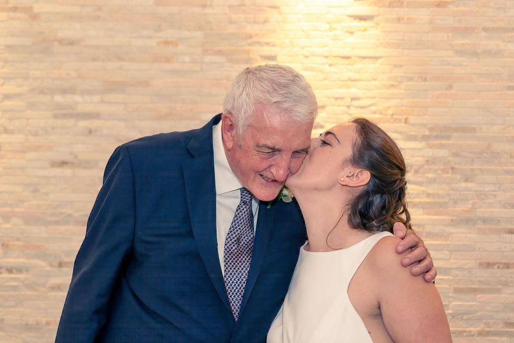 A tender moment between a father and daughter in formal attire sharing an embrace against a warm-lit backdrop.