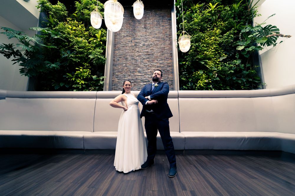 Two people in formal attire pose together on a rooftop deck with hanging lanterns and a stone wall backdrop.