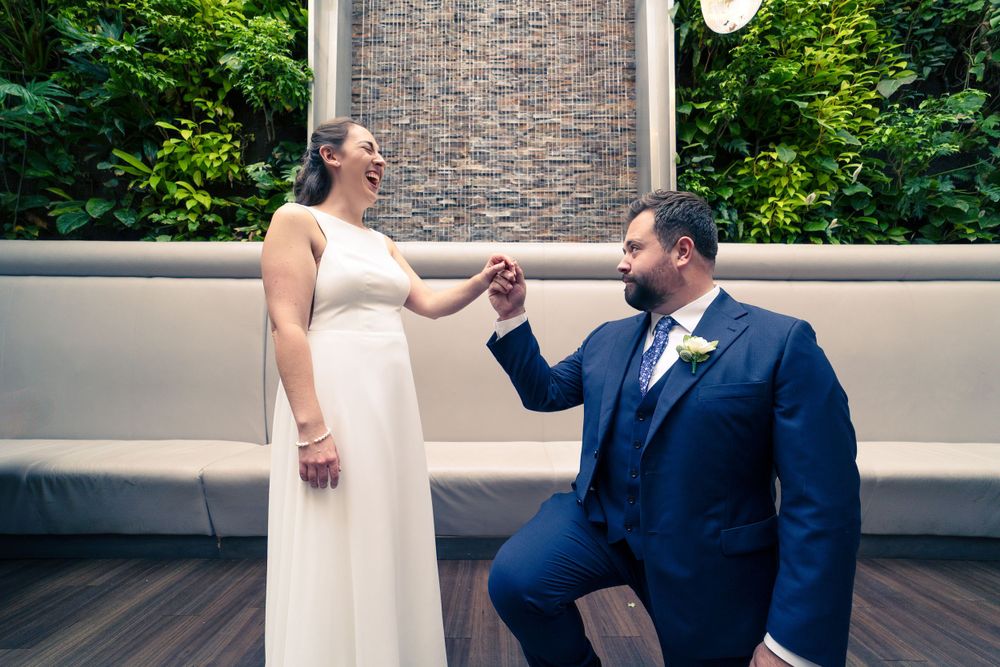 Two people share a sweet moment on a deck with stone wall and greenery, the person in white dress and navy suit.