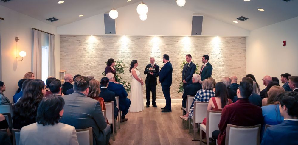 A small indoor wedding ceremony with guests seated in rows watching the couple and officiant at the front of the venue.