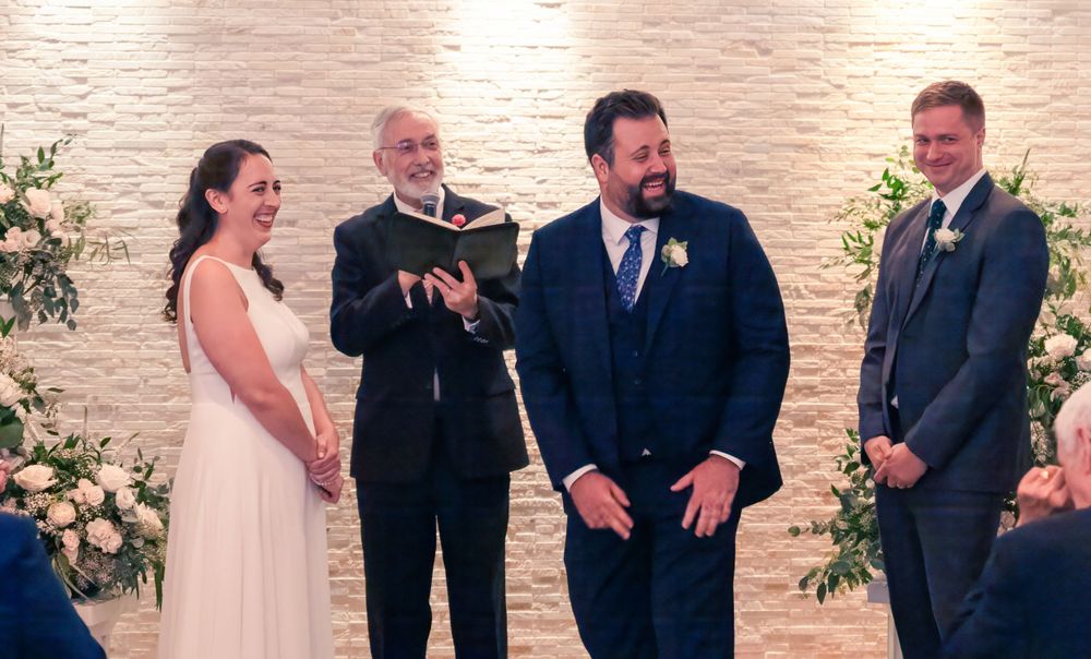 A wedding ceremony takes place with officiant and wedding party at an indoor venue with white brick walls and floral decor.