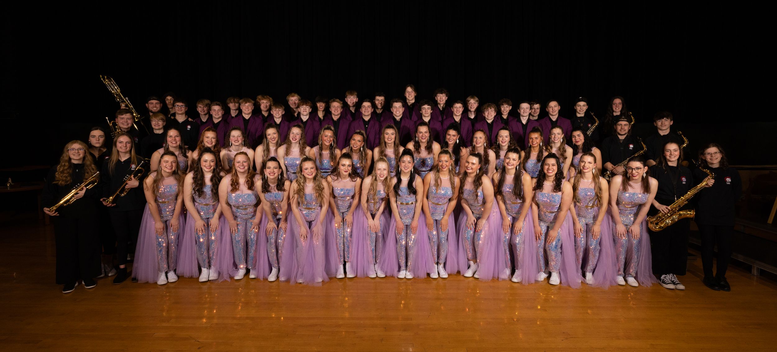 Dance company in lavender dresses lined up for group photo on stage.