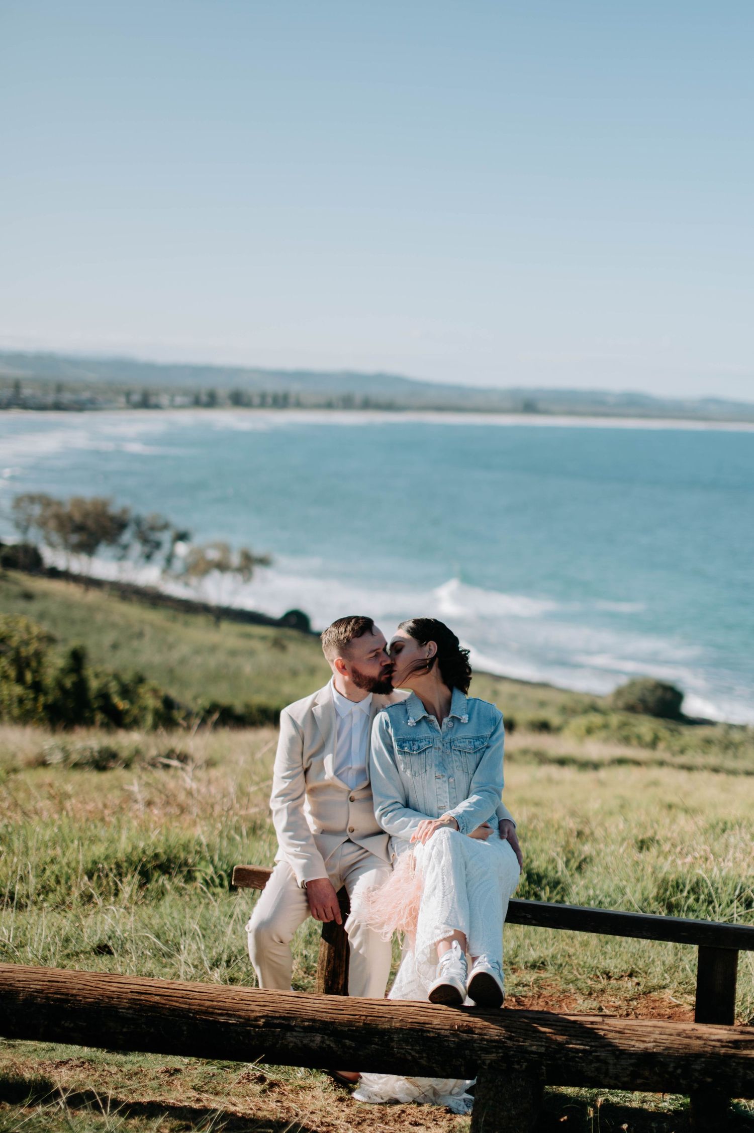 A couple shares a tender moment while sitting on a wooden bench overlooking a scenic coastal view.