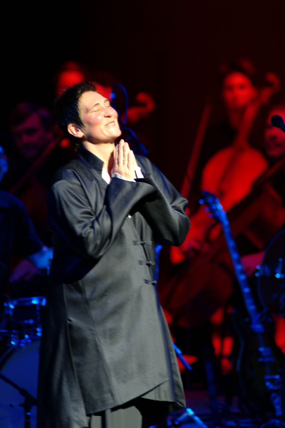 A performer in a long dark coat stands with clasped hands in a prayer pose on stage with dramatic red and blue lighting.