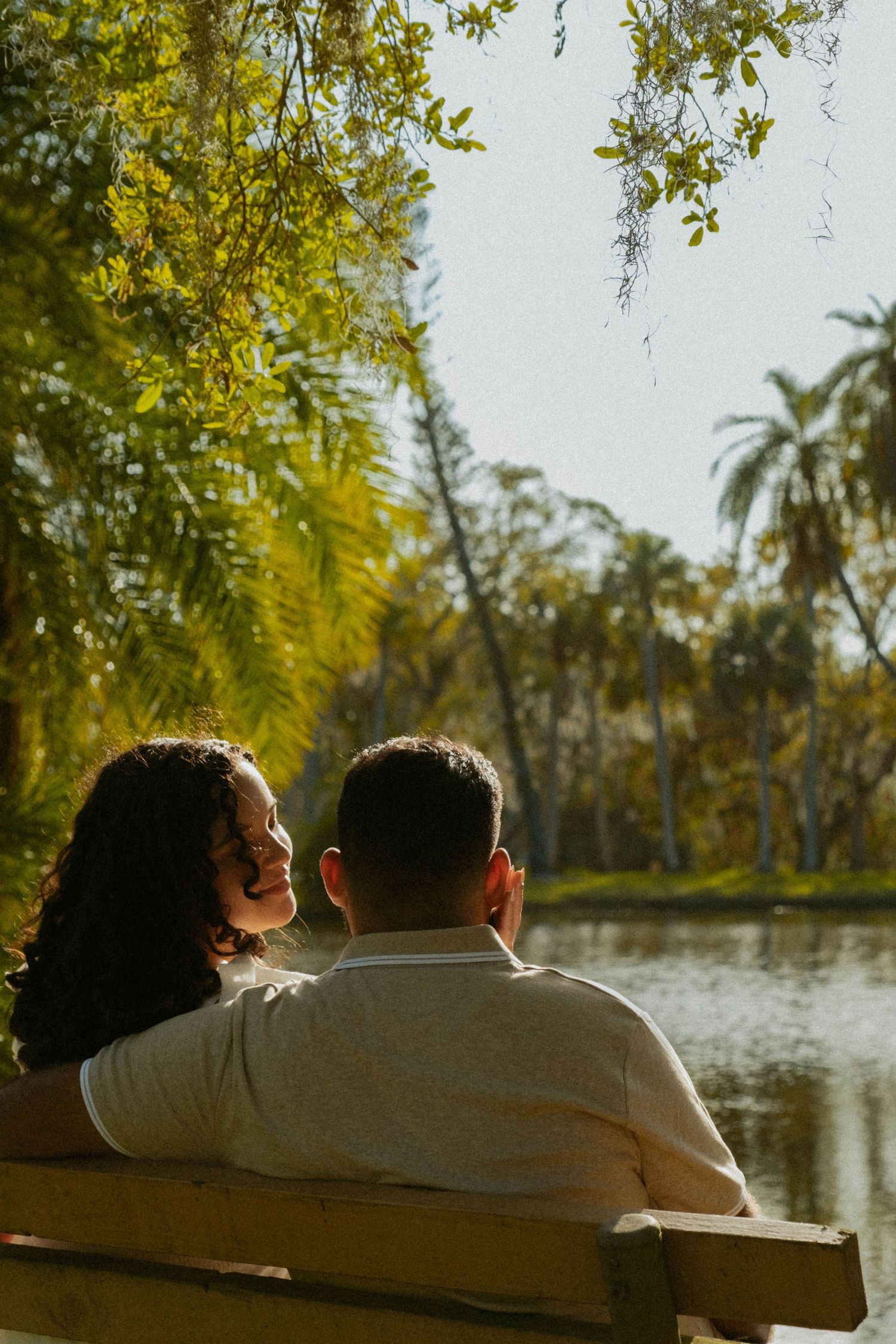 A couple sits together on a bench overlooking a scenic lake surrounded by palm trees.