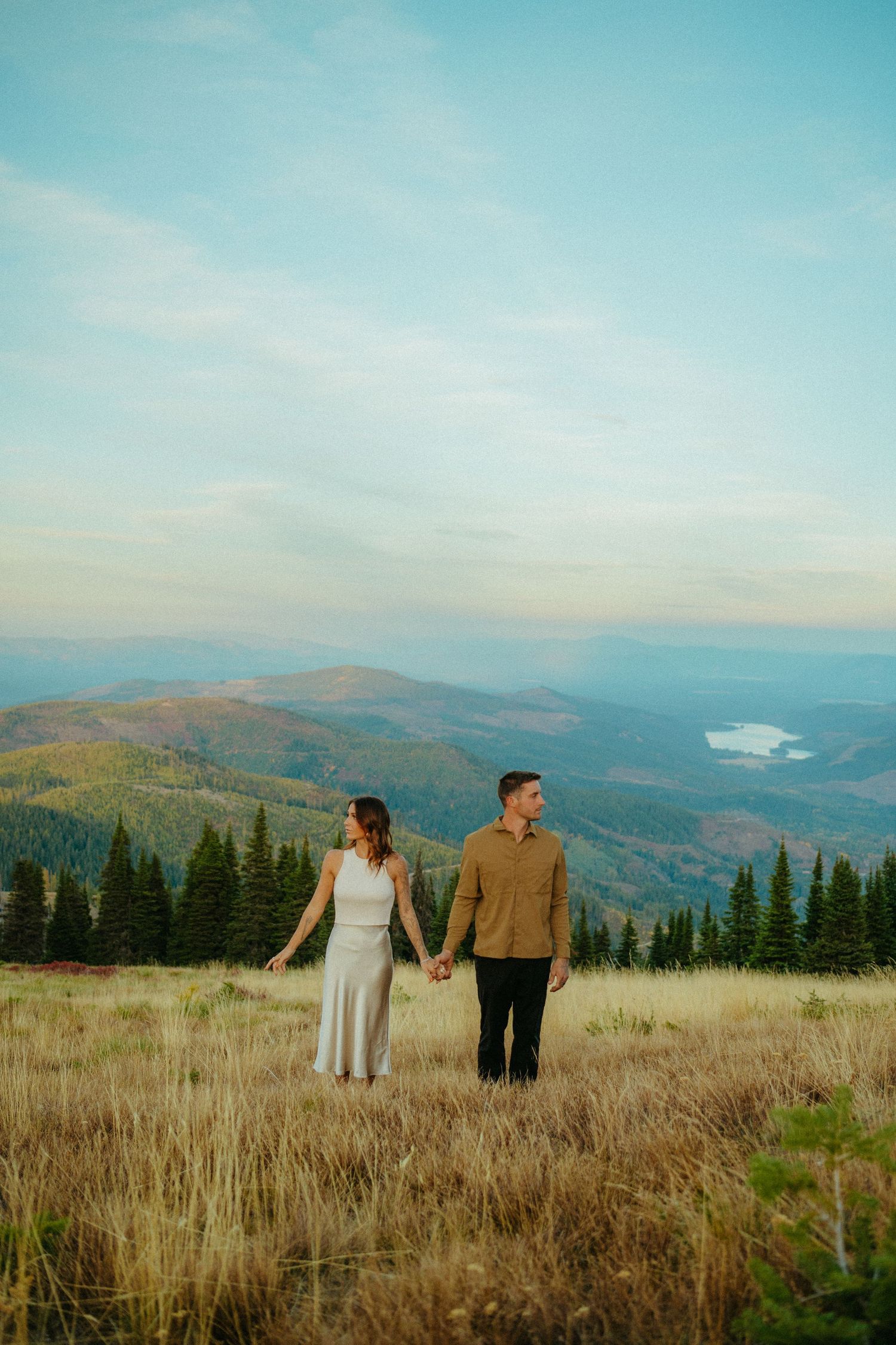 Couple holding hands while standing in mountain meadow with lake view in distance.