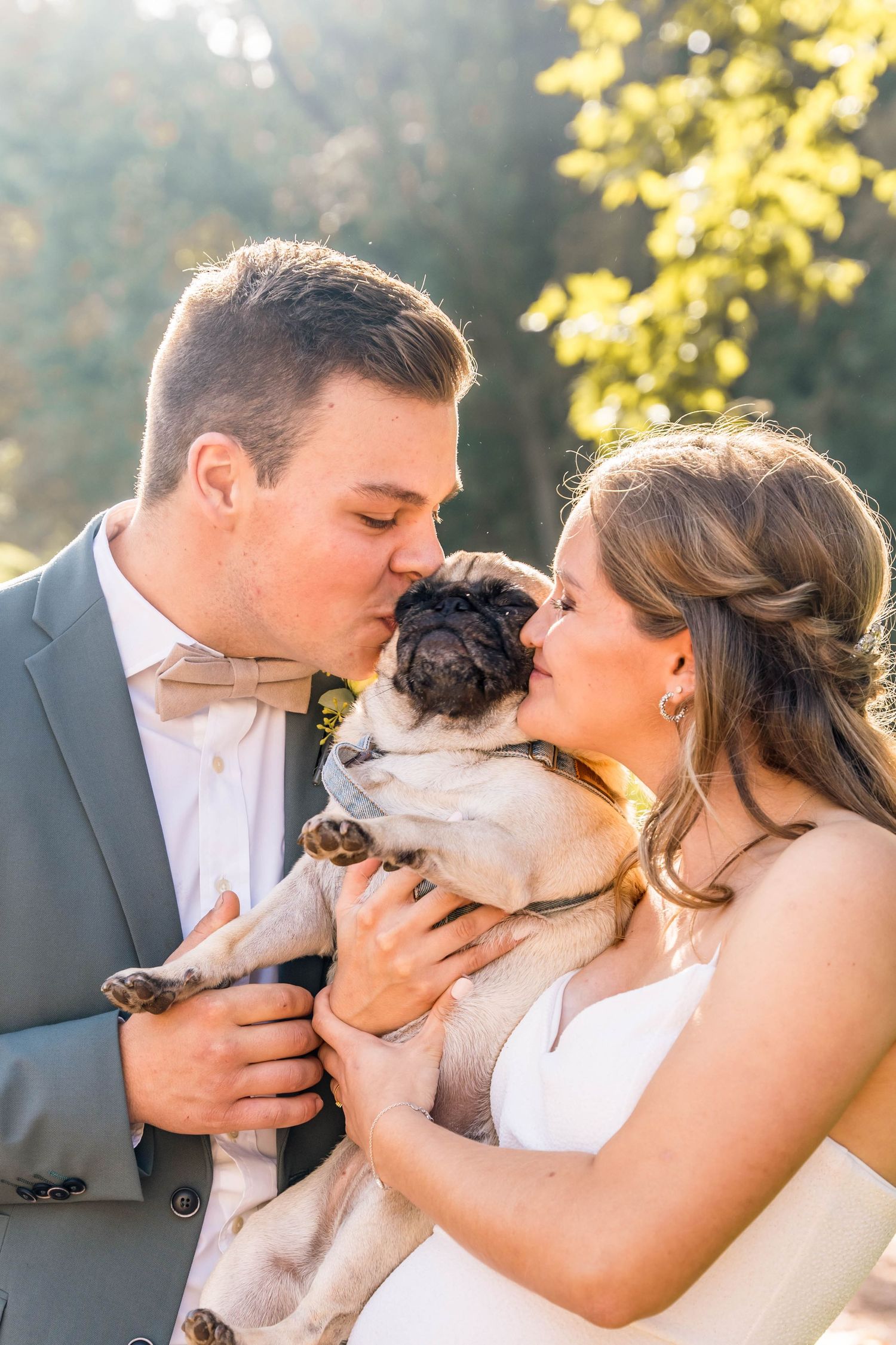A couple in wedding attire holds and kisses their pet pug during an outdoor photo session.