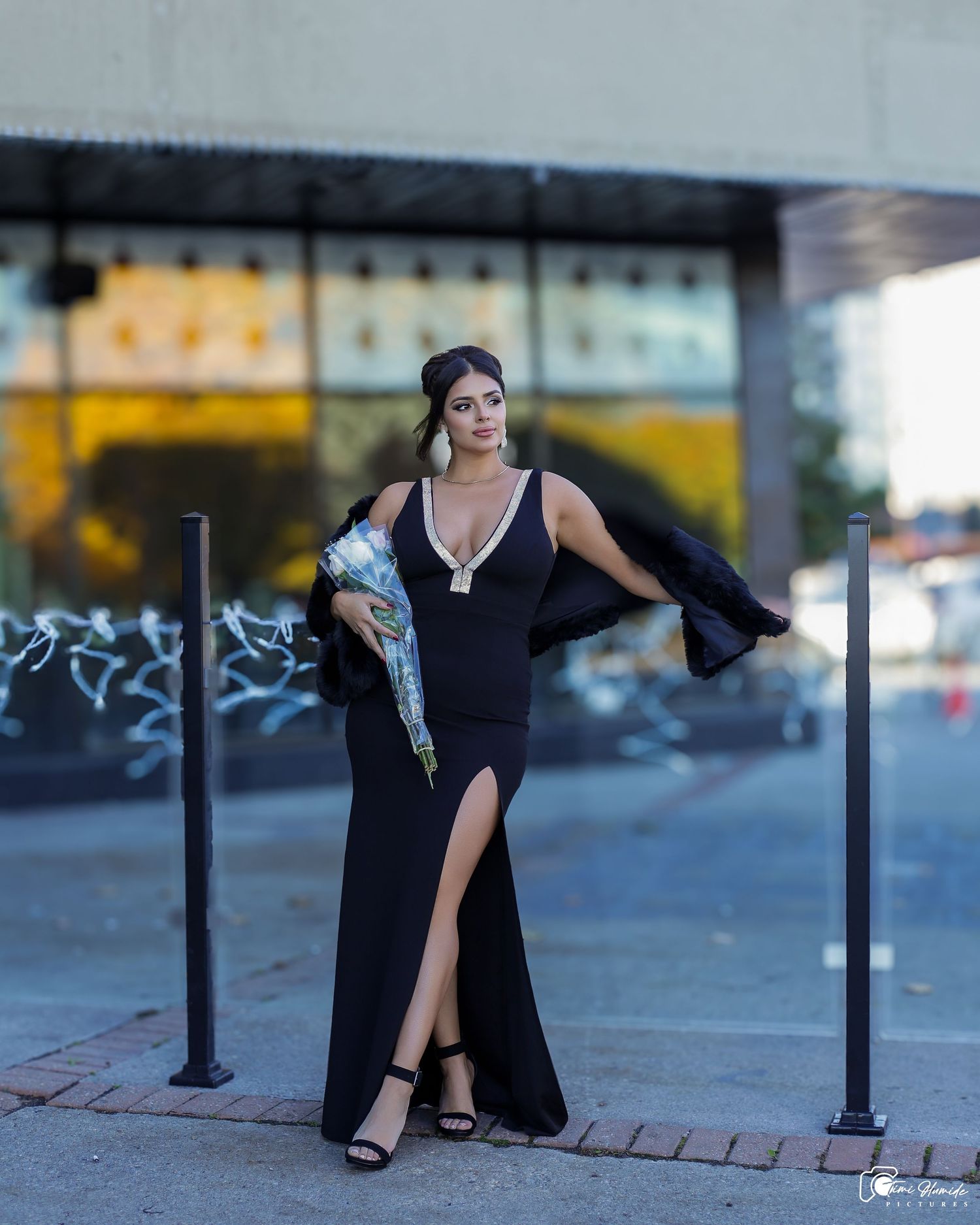 Glamorous fashion photo of someone in a black formal dress with high slit posing on city street.