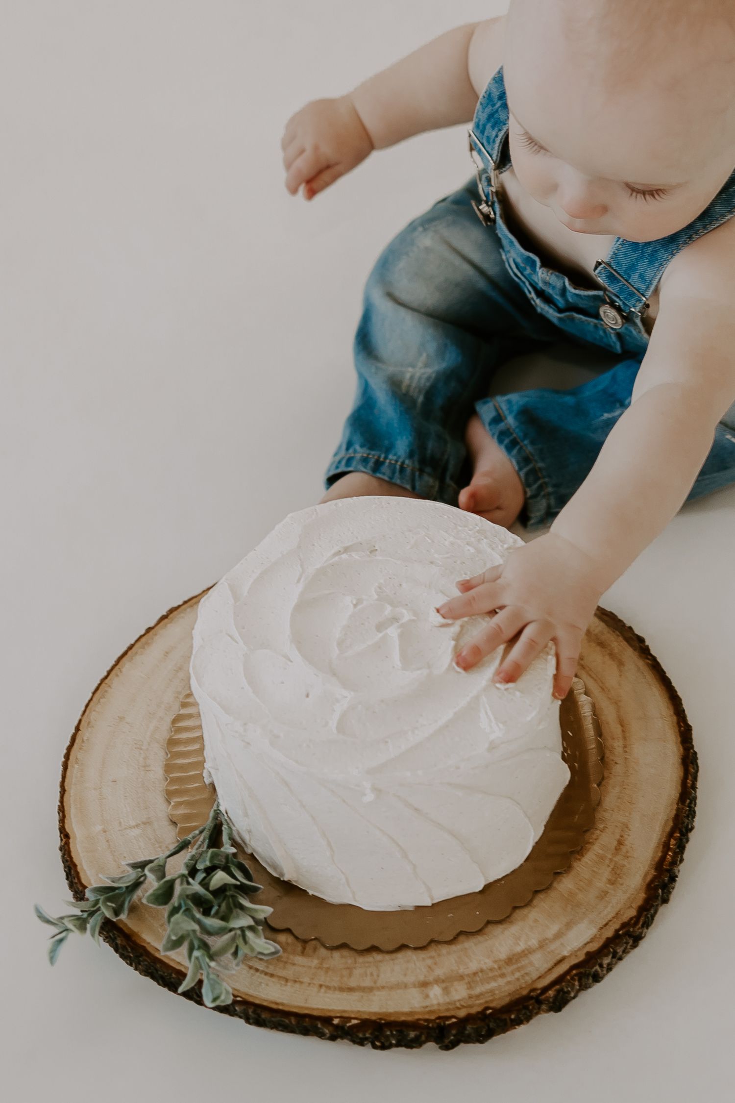 Baby in denim overalls touching a white frosted smash cake on a rustic wood slice.