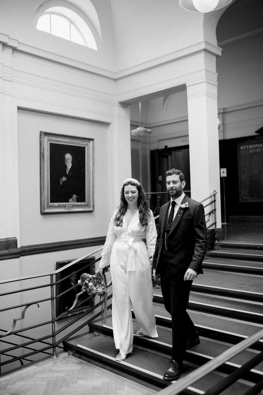 A couple in wedding attire descends an elegant staircase in Stoke Newington Town Hall.