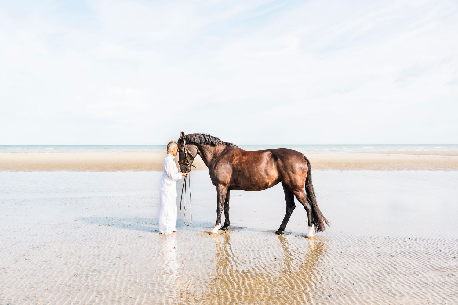 A bay horse and person in flowing white dress stand together on reflective beach waters.