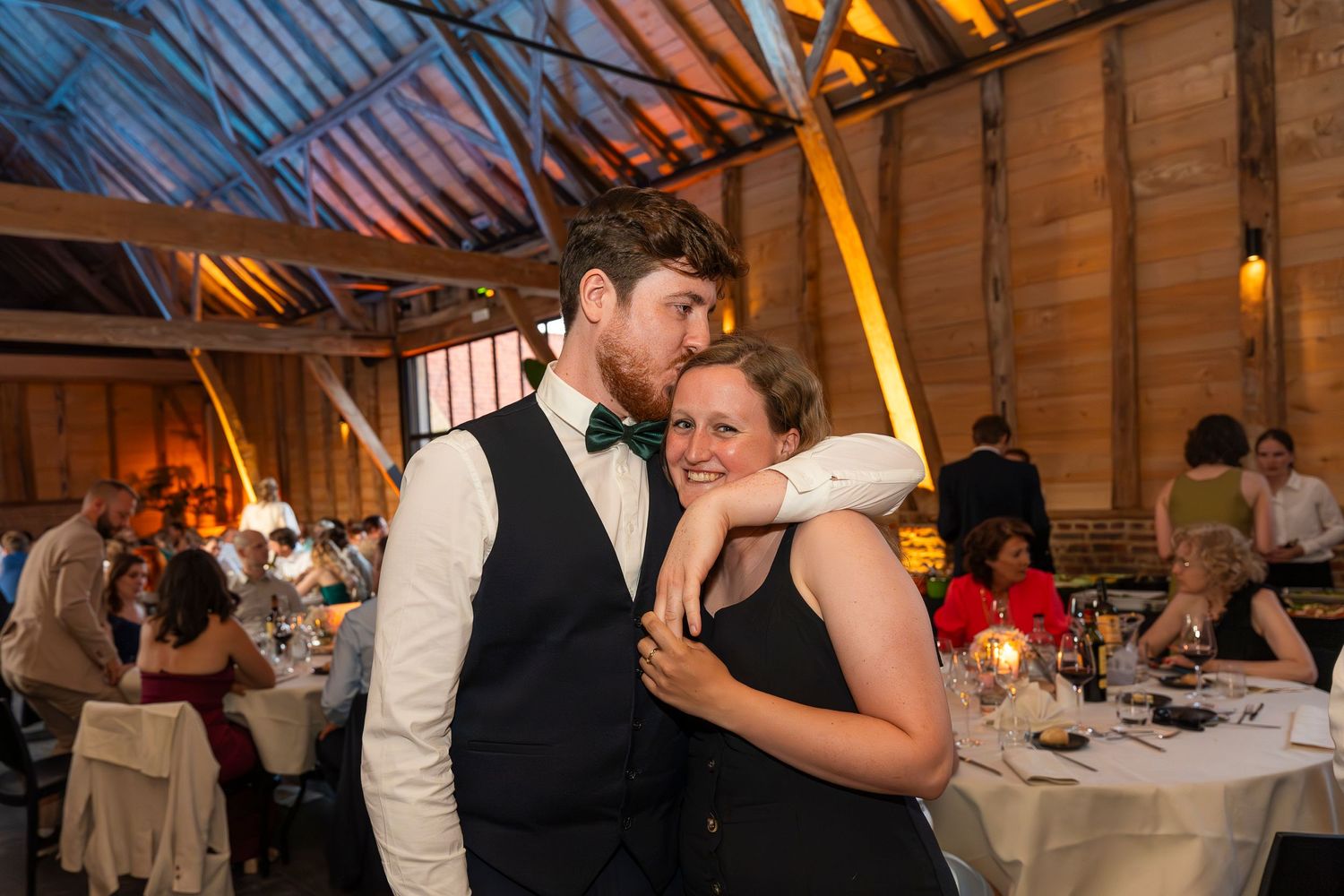 A couple shares an intimate moment during a wedding reception in a rustic barn venue with ambient lighting.