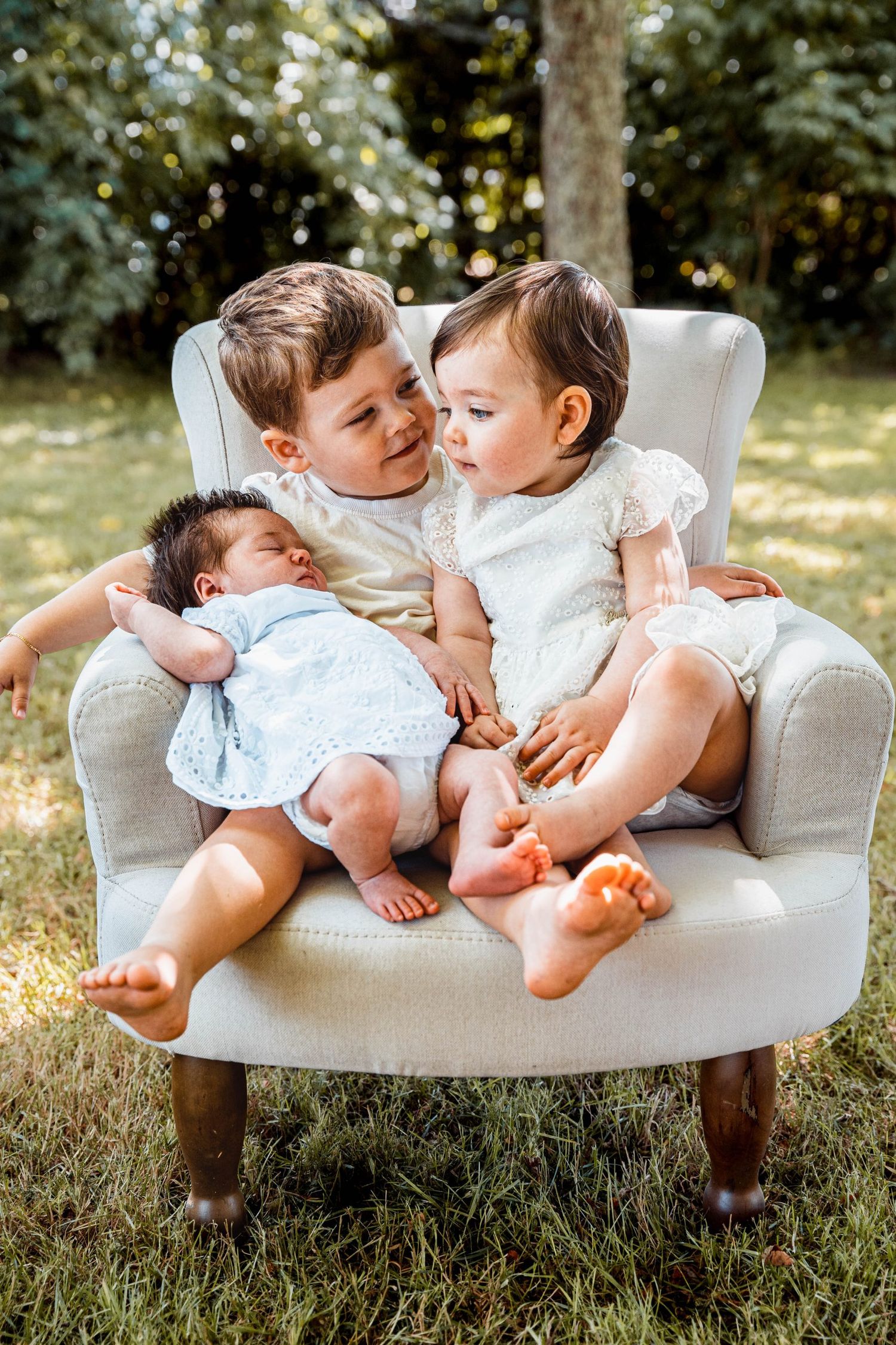 Three children snuggled together on a cozy white armchair outdoors.