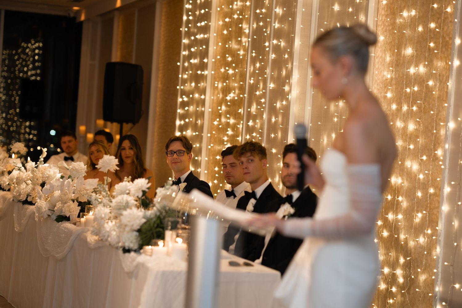 A wedding reception banquet table decorated with white florals and glowing string lights in the background.
