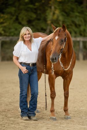 Person in jeans and white shirt poses with brown horse in sandy arena.
