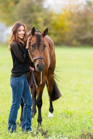 Person in jeans stands with a brown horse in a grassy field on a sunny day.