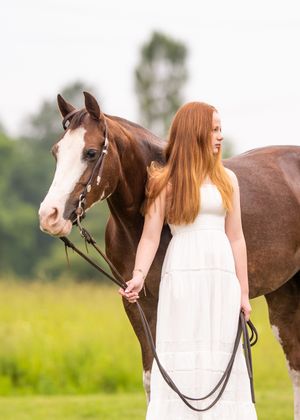 Person in white dress poses with brown horse in sunny field setting.