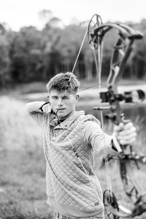 A person in a quilted sweater practices archery with a bow and arrow at a lakeside outdoor setting.