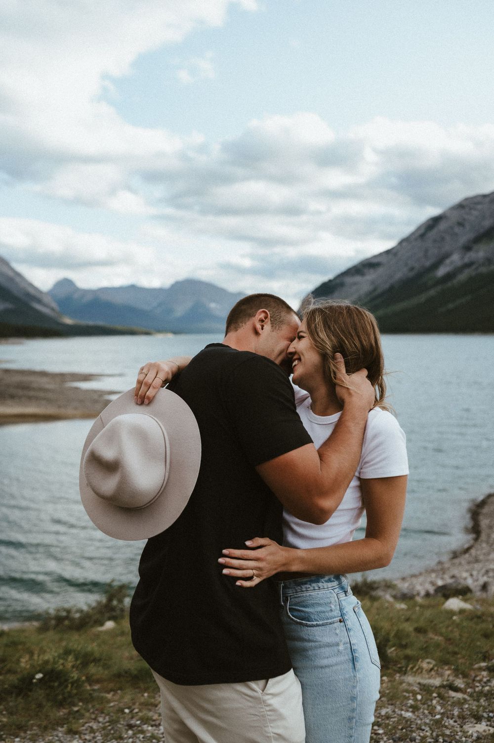 Romantic couple laughing together by a mountain lake while holding a wide-brimmed hat.