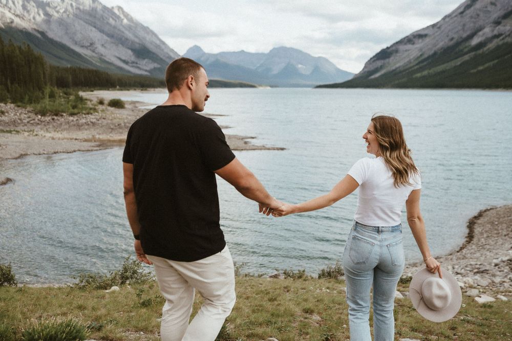 Couple walking hand in hand along a rocky lakeshore with mountains in the background.