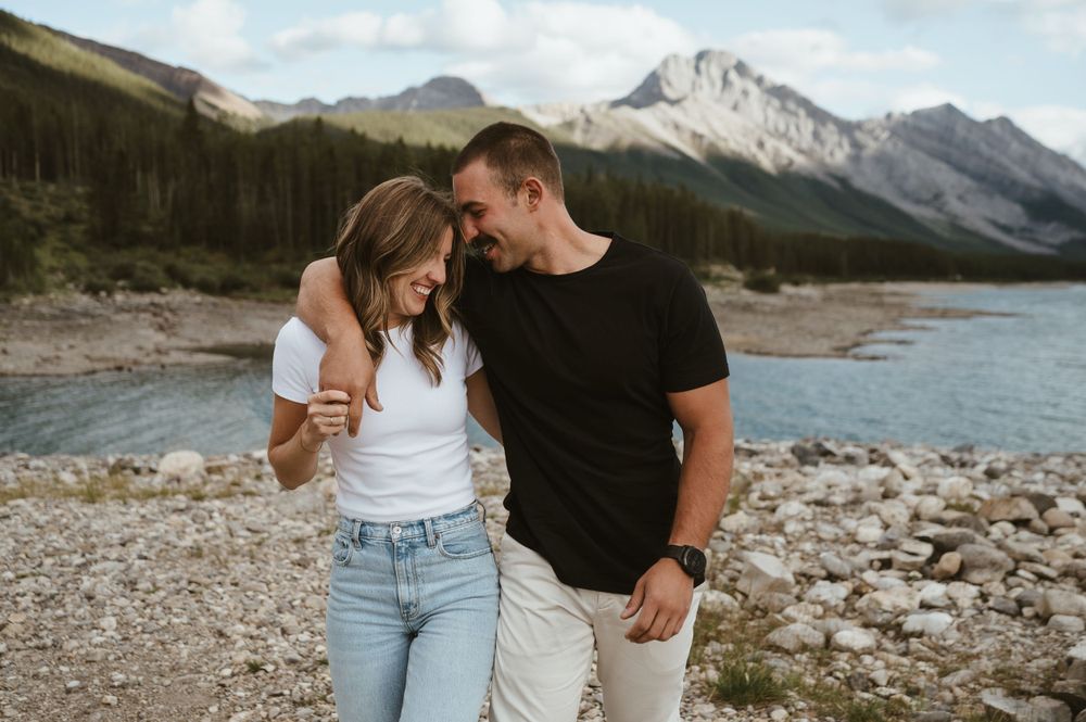 Couple sharing an intimate moment on rocky shoreline with mountain peaks in background.
