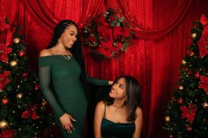 Two people in elegant dresses pose against a red curtain backdrop with Christmas decorations.