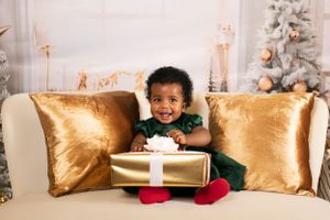 A small child in green sits on a gold couch holding a large wrapped present during Christmas.