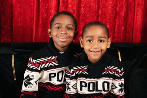 Two children in matching Polo holiday sweaters smile while seated against a red backdrop.