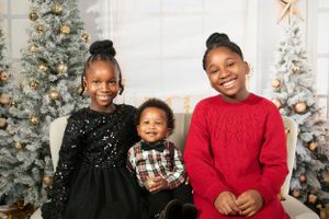 Three siblings pose for a Christmas photo in front of decorated trees wearing festive holiday outfits.