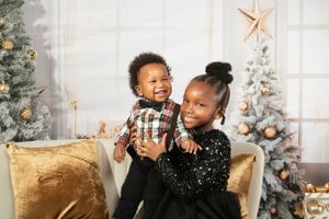 Two children share a joyful moment on gold chairs with Christmas trees in the background.