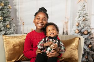 Two siblings in festive holiday wear share a happy moment in front of decorated Christmas trees.