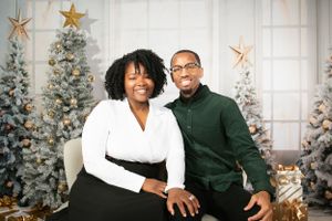 Couple in white and green outfits pose in front of decorated Christmas trees with gold star ornaments.