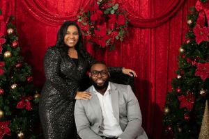 Elegant couple poses against red backdrop with Christmas decorations and wreaths.