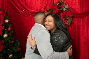 Couple embracing and smiling against red curtain backdrop with holiday decorations.