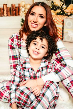 Two people wearing matching plaid Christmas pajamas smile while sitting on a white fur rug with holiday decorations.