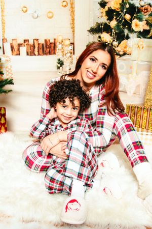 Two people in coordinating red plaid sleepwear embrace while sitting on white fur near a Christmas tree.