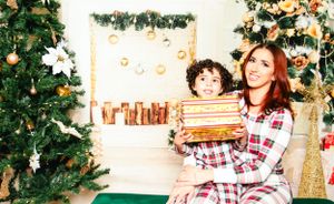 Two people in matching plaid pajamas sit by a Christmas tree with wrapped presents.