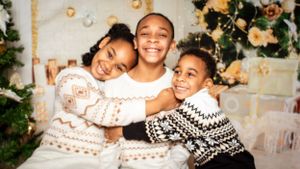 Three siblings wearing cozy winter sweaters laugh and hug during a Christmas photoshoot.
