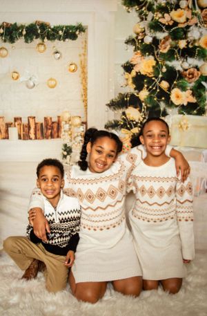 Three siblings wearing matching cream and brown patterned sweaters pose in front of a decorated Christmas tree.