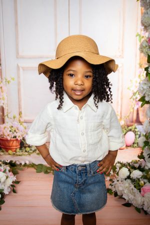 Child in white shirt and straw hat poses in front of floral garden backdrop.