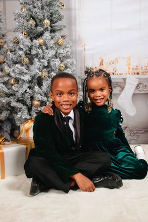 Siblings in velvet holiday outfits sitting together by decorated Christmas tree.