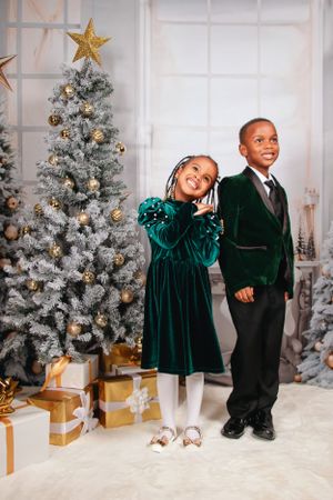 Children in formal attire posing by decorated Christmas tree with gold wrapped presents.